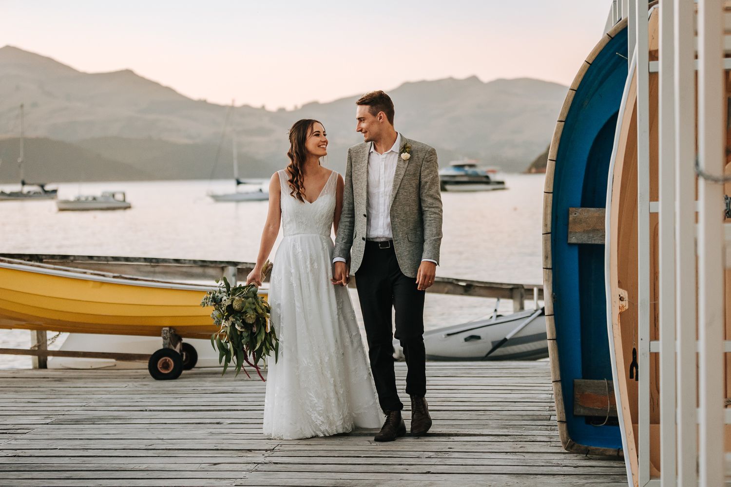 wedding bride and groom holding hands on jetty with boats in background at sunset in akaroa