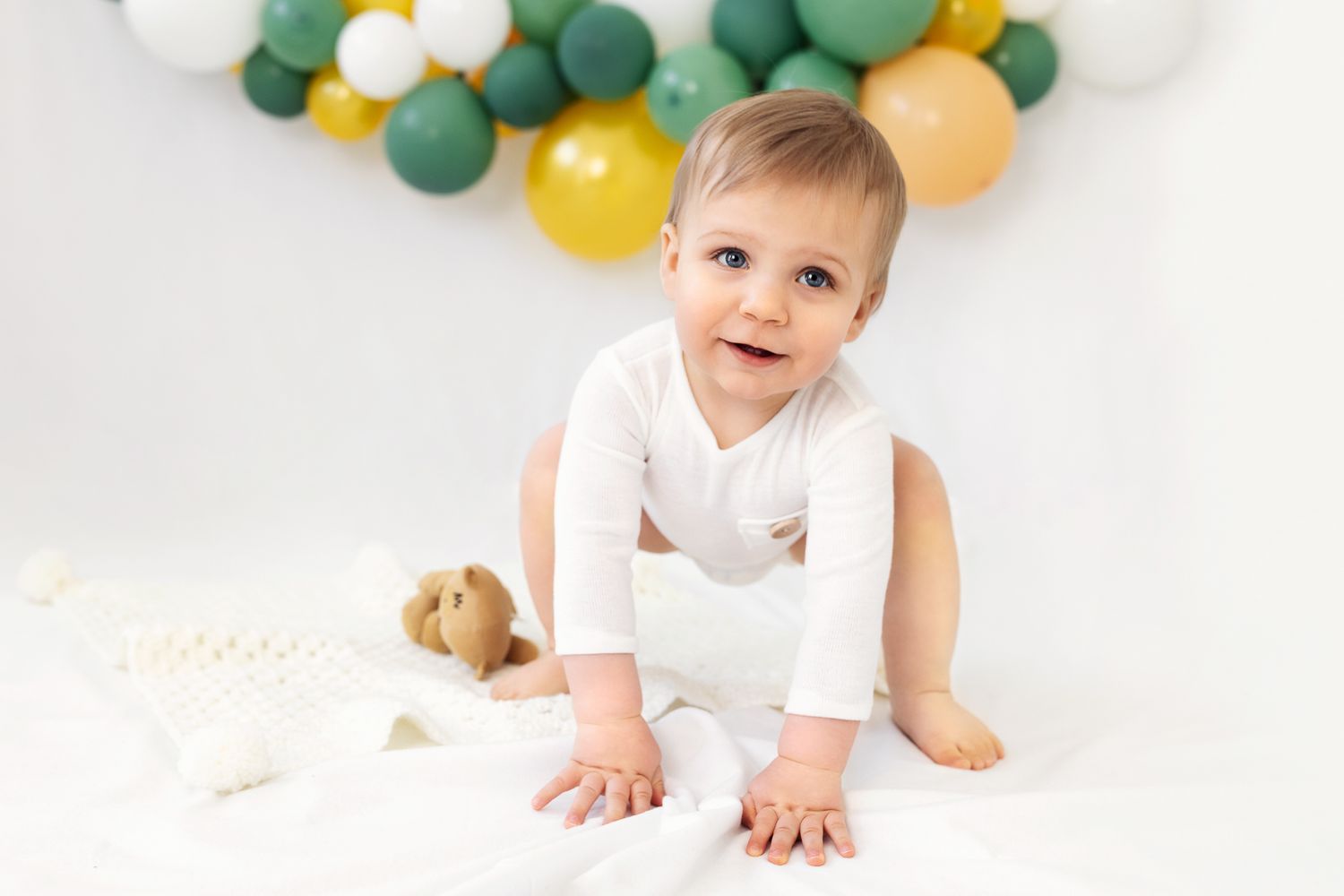 Infant in white onesie playing on floor with festive balloon arch in background.
