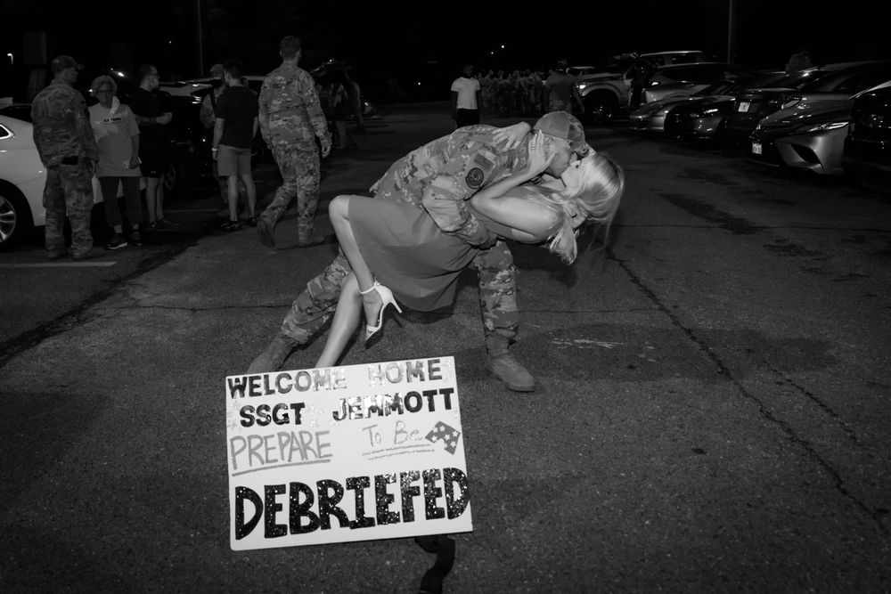 Black and white image of soldier lifting woman during emotional military homecoming reunion outdoors.