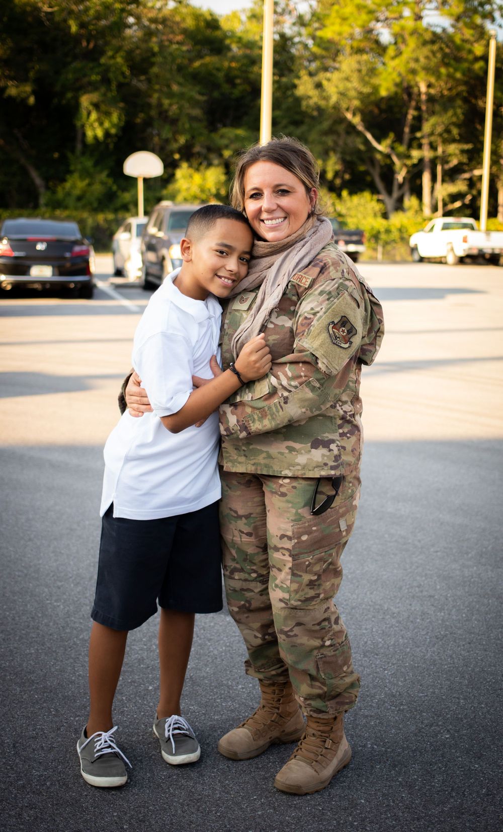 Female soldier in camouflage uniform hugs smiling young boy in parking lot outdoors.