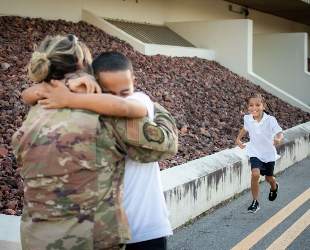 Military soldier in uniform embraces family member while child runs toward them for reunion hug.