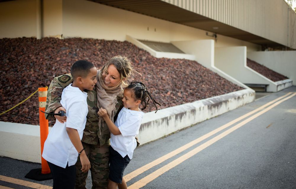 Female soldier in uniform shares joyful embrace with two children during homecoming reunion.