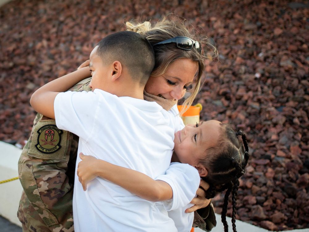 Military mother emotionally embraces two children in a heartfelt homecoming reunion outdoors.
