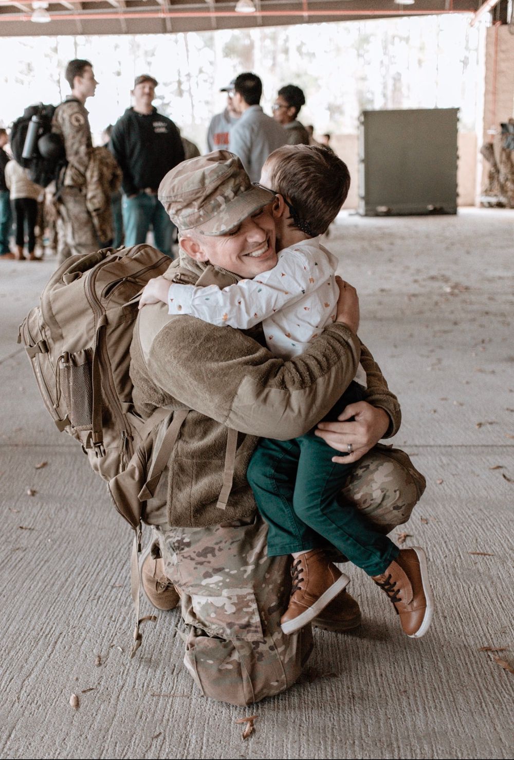 Soldier kneels to hug excited child during emotional military homecoming reunion indoors.