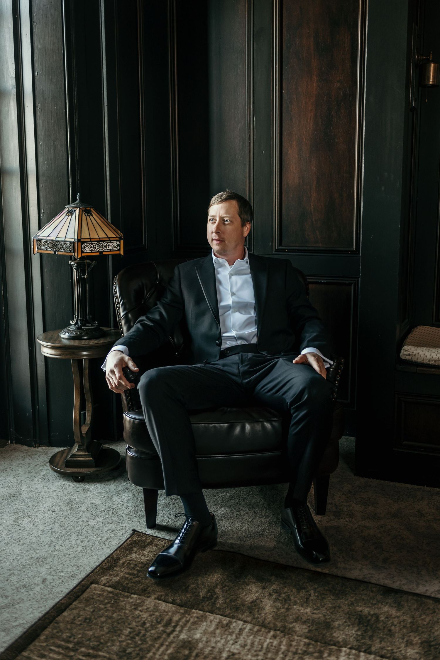 Man in dark suit sits contemplatively in leather chair against wood-paneled wall.