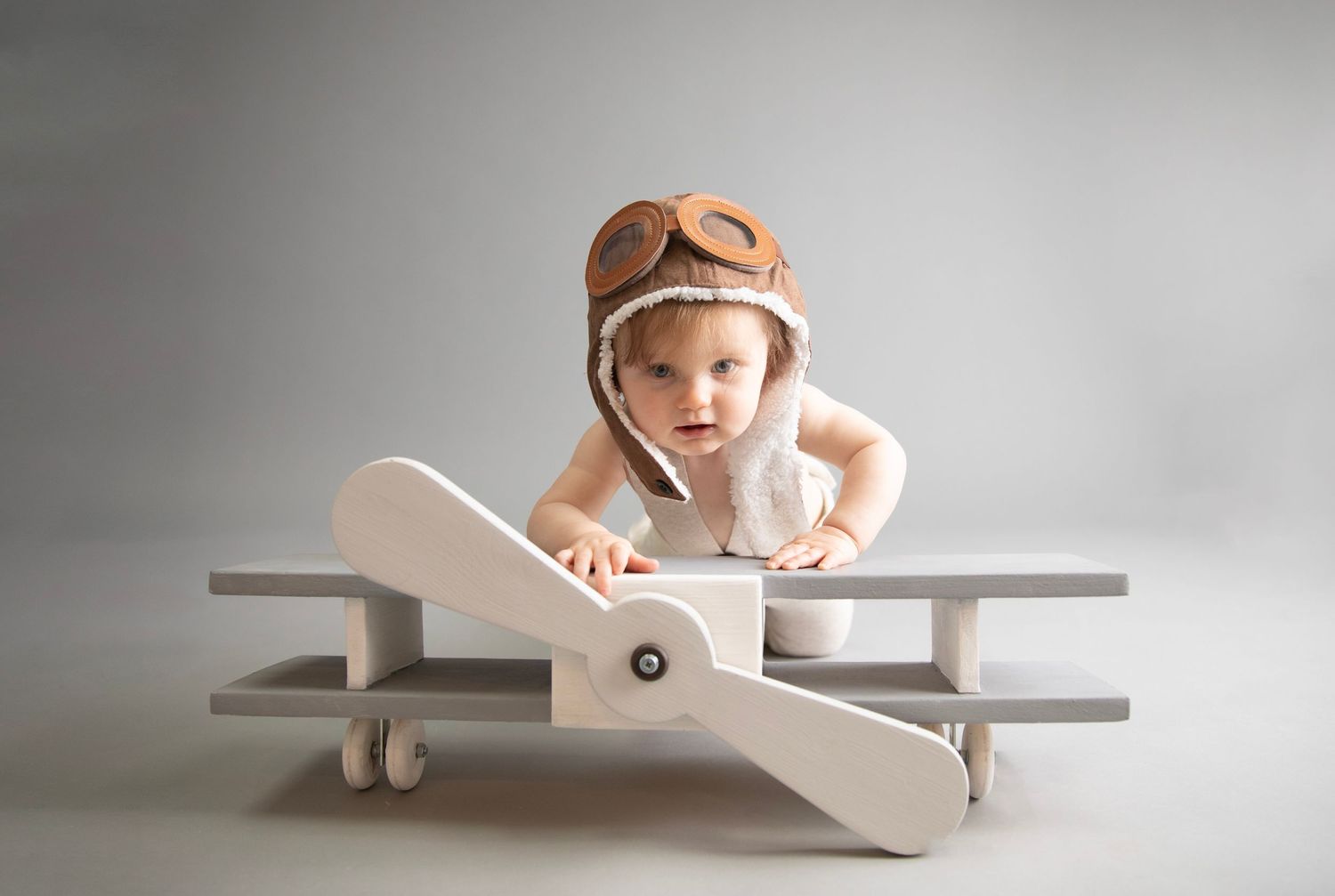 Cute portrait sitter session. child on wooden aeroplane prop, wearing pilot hat & goggles. grey backdrop