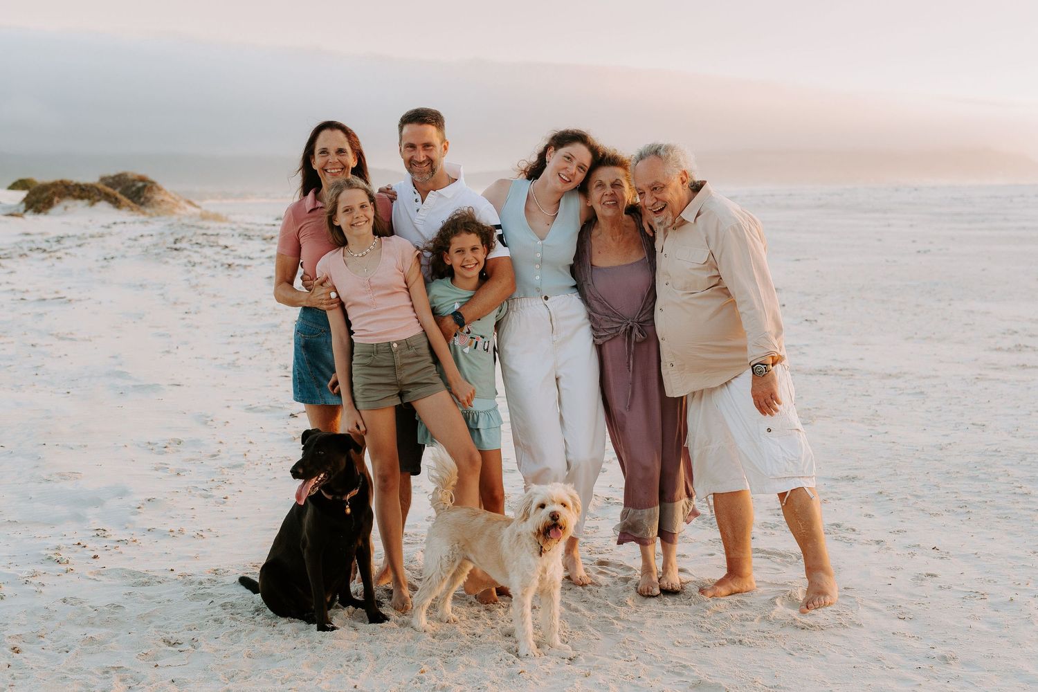 A cheerful group poses with their dogs on a sandy beach at sunset.
