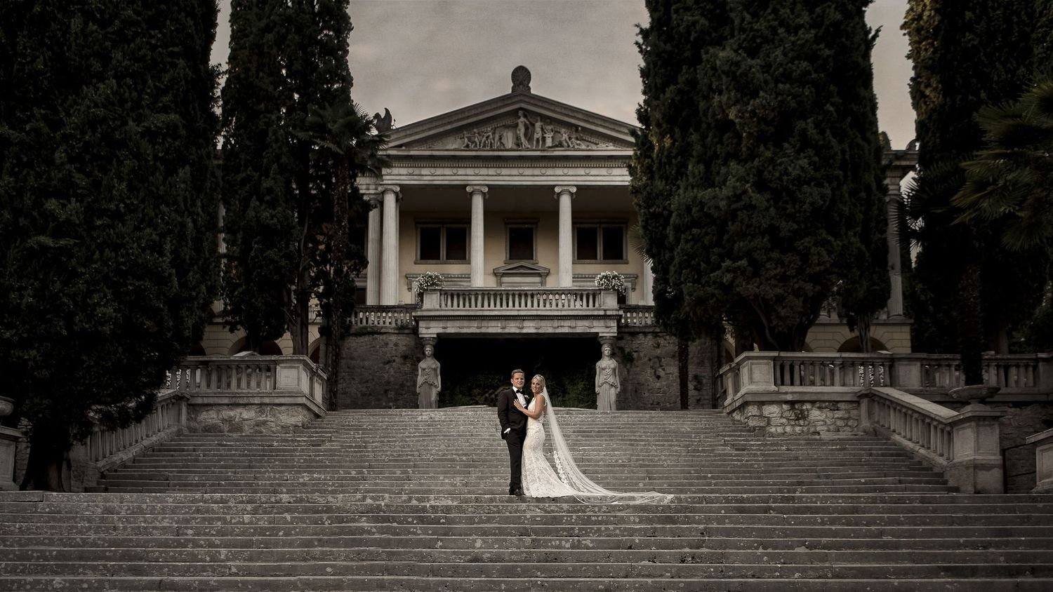 Elegant white neoclassical building with columns and stone steps surrounded by trees in a garden setting.
