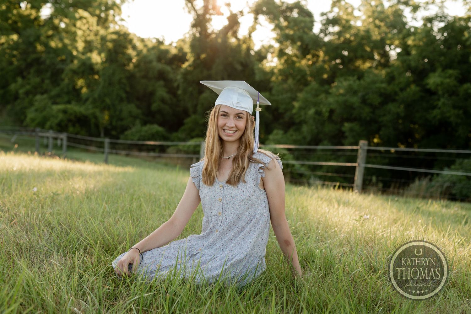 Graduate in white cap and grey dress poses in rustic field setting during golden hour.