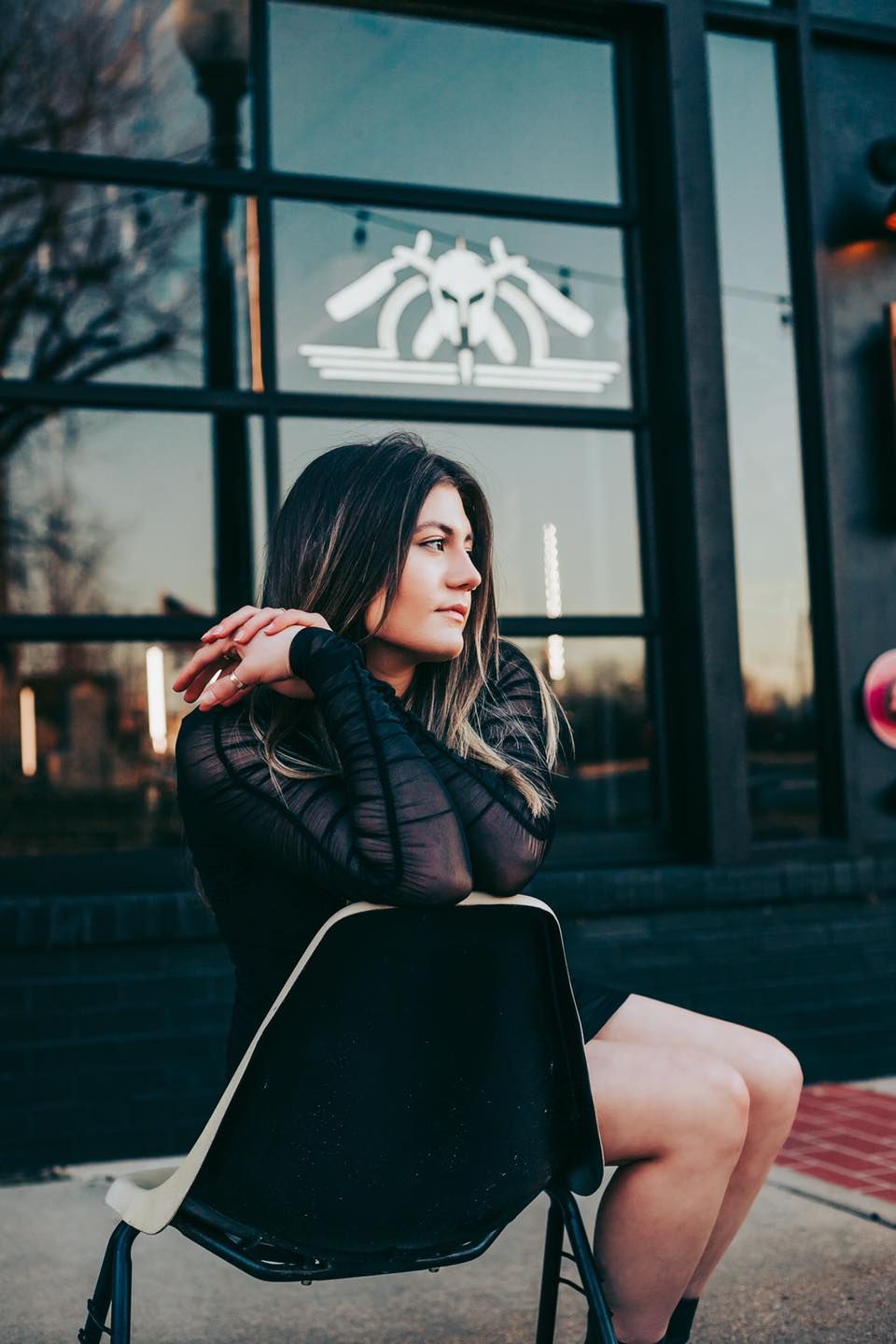 A moody outdoor portrait of a woman in black sitting in a chair at sunset with architectural details in background.