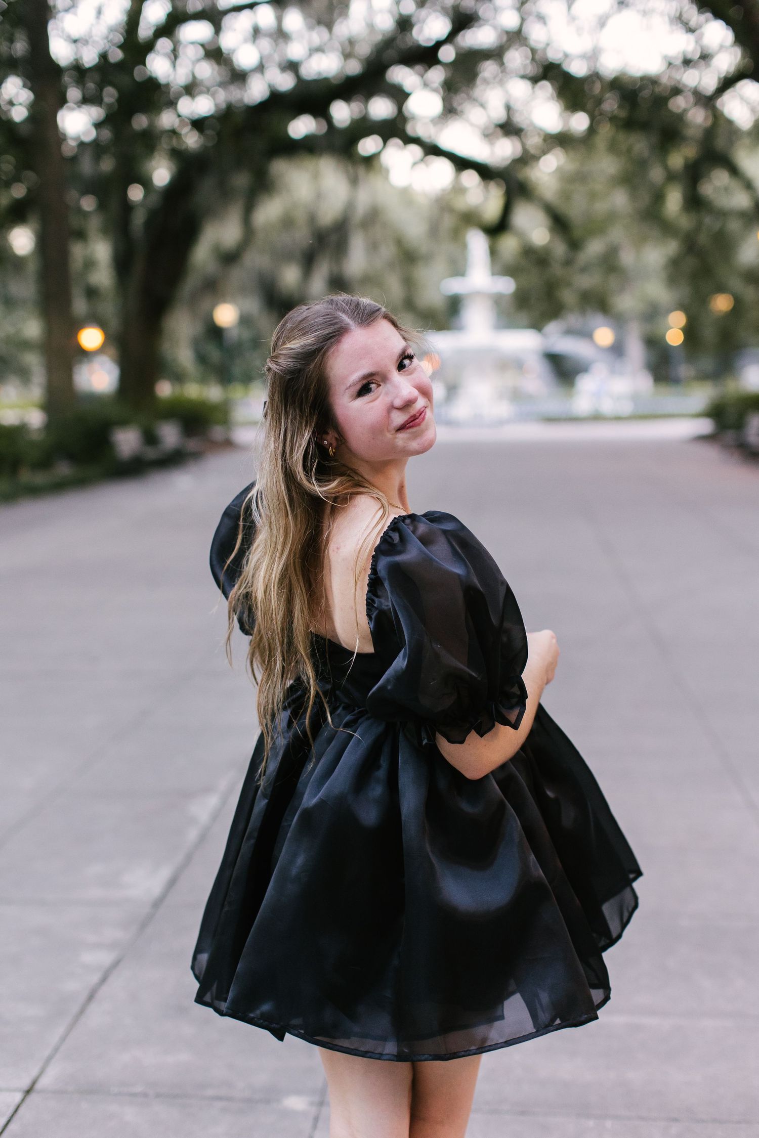 Someone in a black dress poses playfully on a tree-lined path with a fountain visible behind.