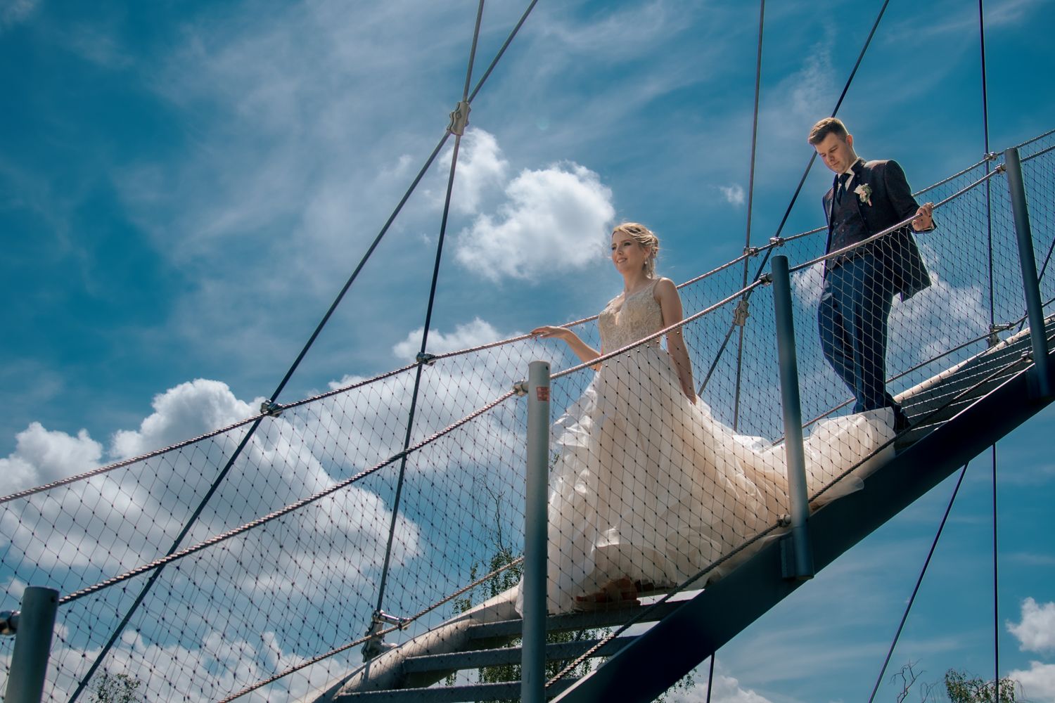 Wedding couple crossing suspension bridge against blue sky with clouds.