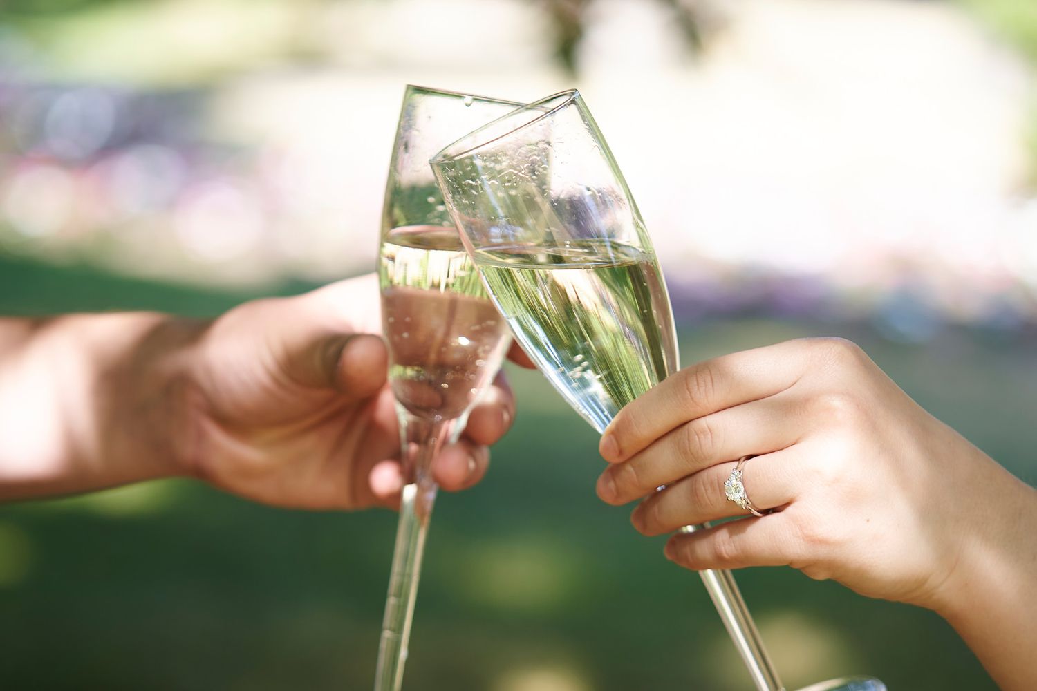 Two hands clinking champagne glasses together in celebration of an engagement showing off engagement ring.