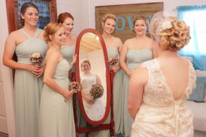 Bridesmaids in sage green dresses gather around mirror during wedding preparations.