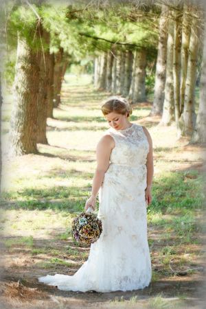 Bride in lace gown holds rustic bouquet while standing in serene forest setting.