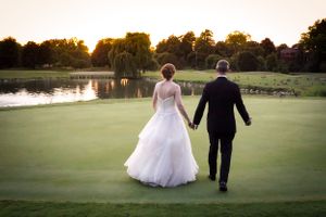 A sunset wedding photo of a couple walking hand in hand on a golf course near a pond.