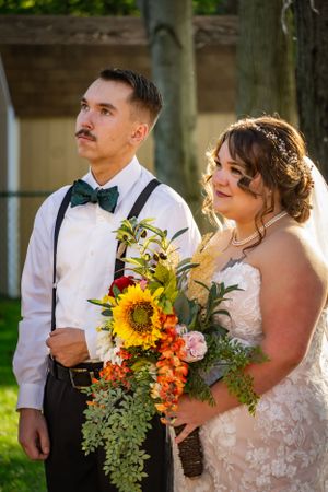 Wedding couple poses outdoors with bright sunflower bouquet and groom wearing suspenders and bow tie.