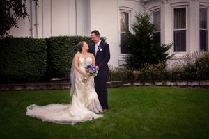 Wedding couple poses on lawn in front of historic building with elegant landscaping.