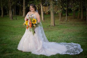 Bride in lace dress with long train holds vibrant sunflower bouquet in outdoor garden setting.
