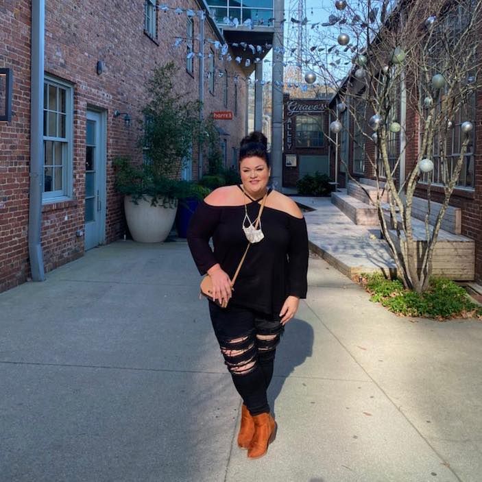 Plus-size woman in black off-shoulder top and ripped jeans posing in urban brick alley.