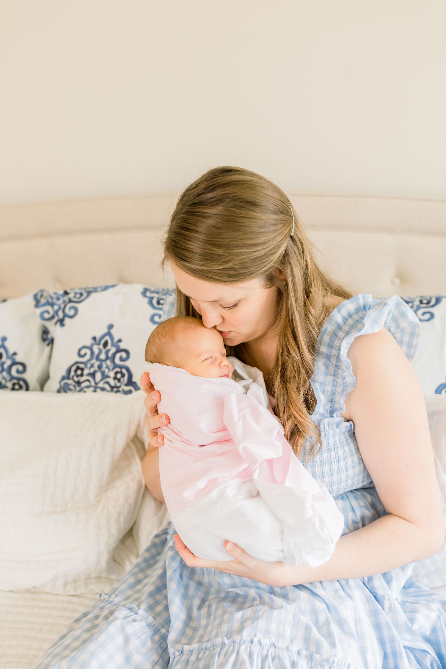 Mother in blue dress tenderly kisses newborn baby while sitting on bed.