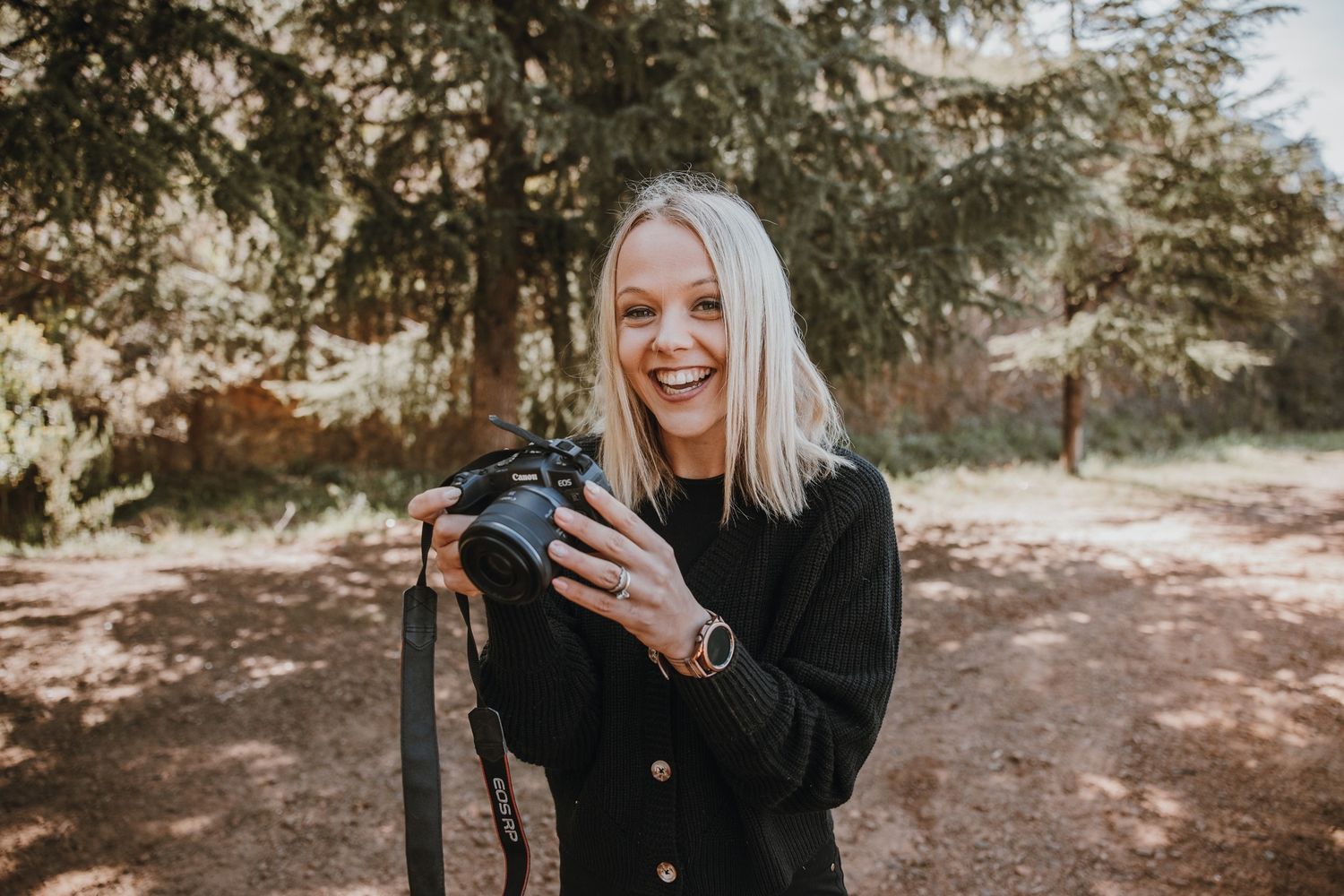 Outdoor photographer in black coat smiles while holding camera equipment on autumn forest path.