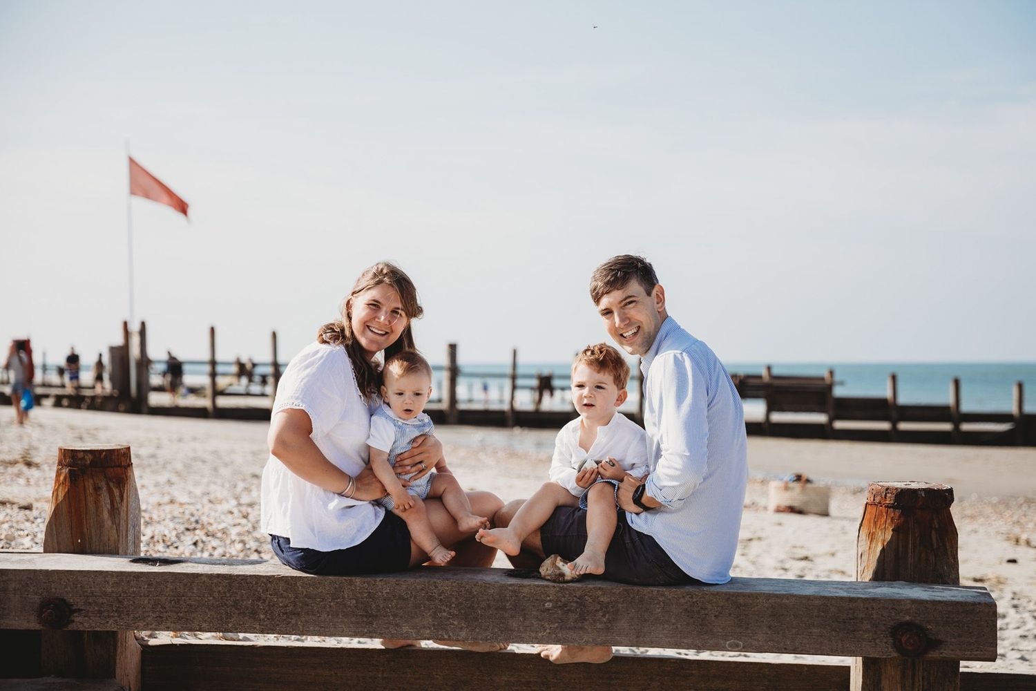 Family relaxing together on wooden beach bench with ocean and pier in background.