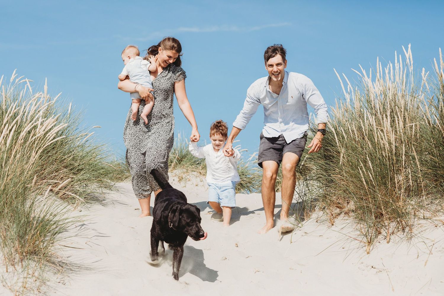 Family running on beach dunes with their black dog on a sunny day.
