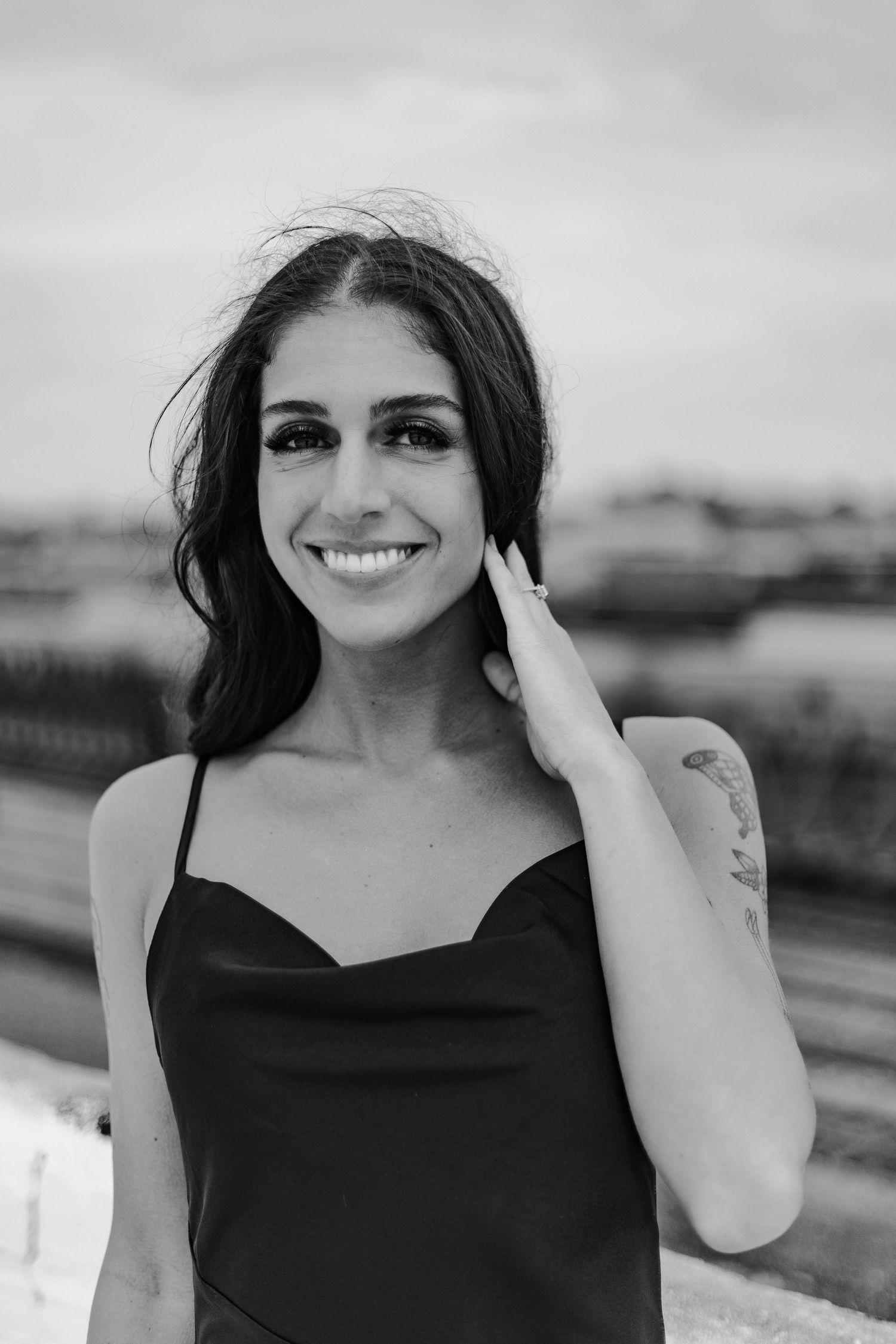 Smiling brunette woman in black dress with arm tattoo poses outdoors in black and white portrait photo.
