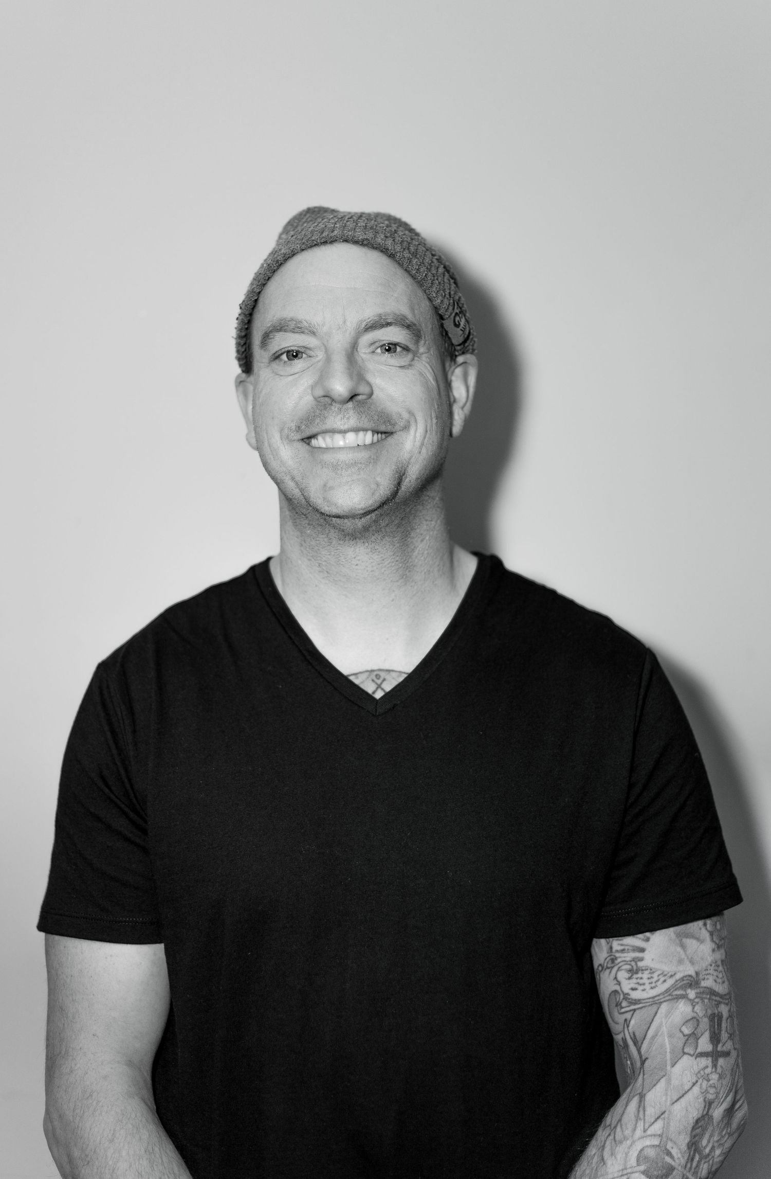 Smiling tattooed man in a black v-neck t-shirt and grey beanie in black and white photo.