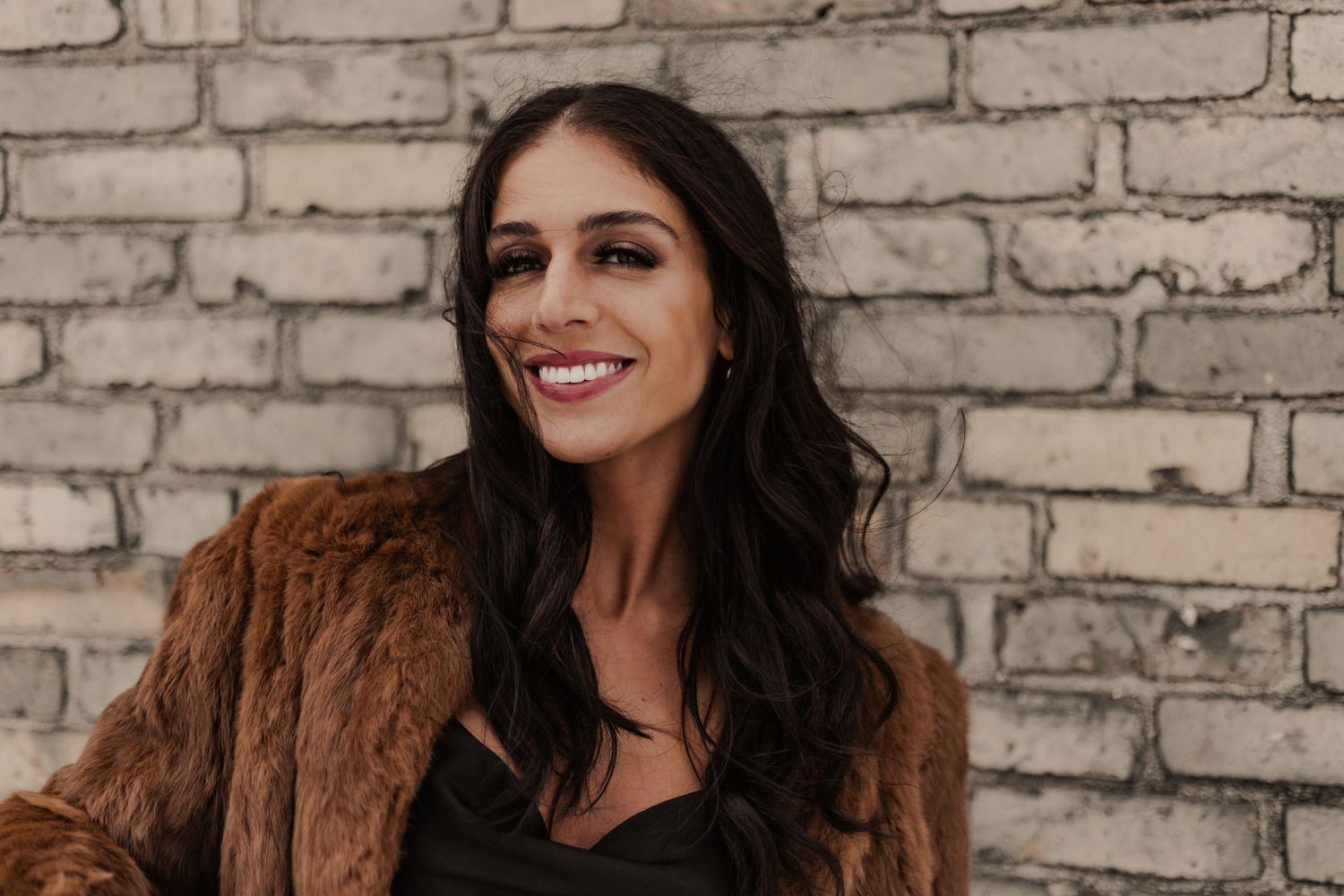 Brunette woman smiling brightly in brown fur coat against white brick wall in warm portrait photo.