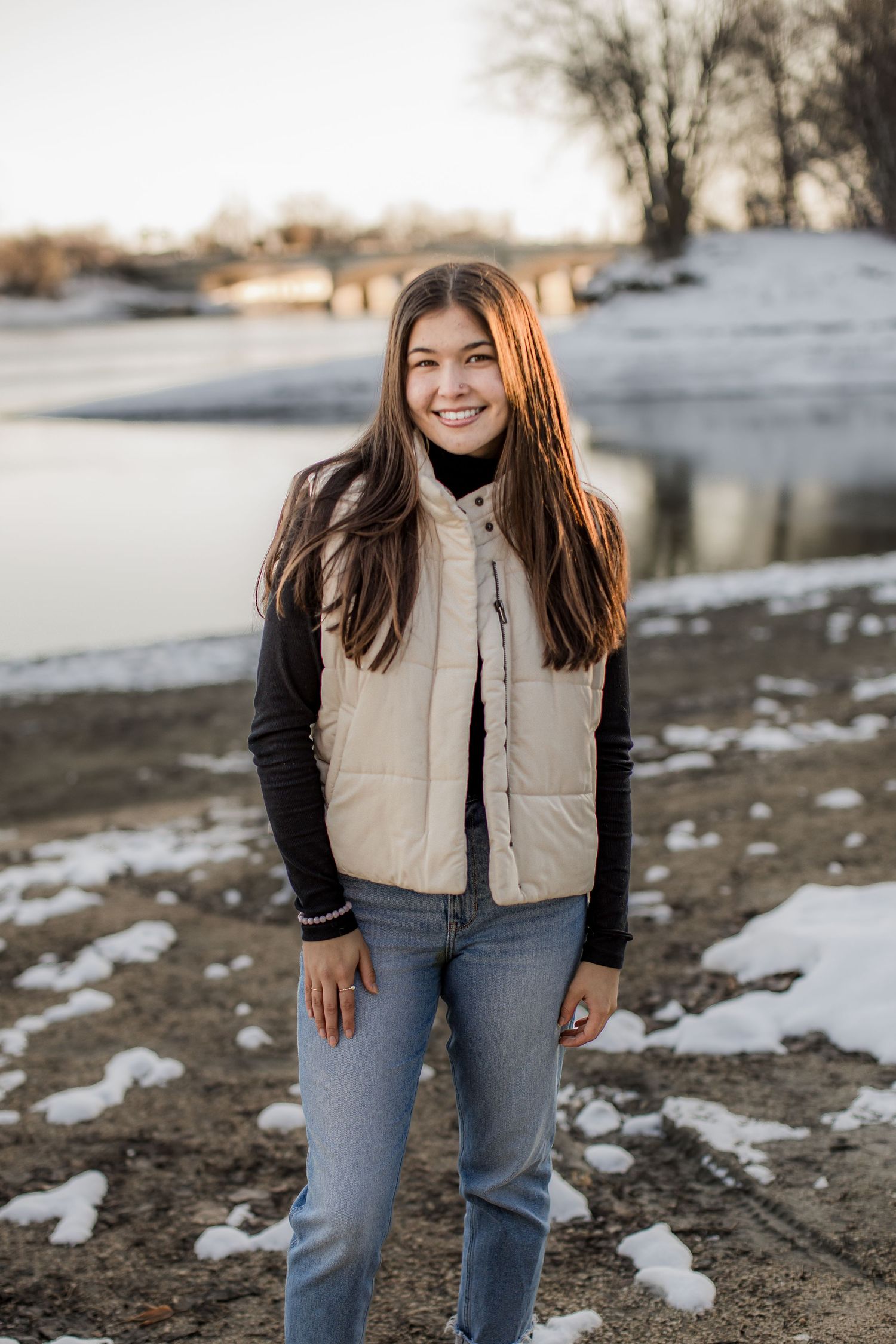 Smiling teenage girl in beige puffer vest and jeans standing near a snowy riverbank at golden hour.