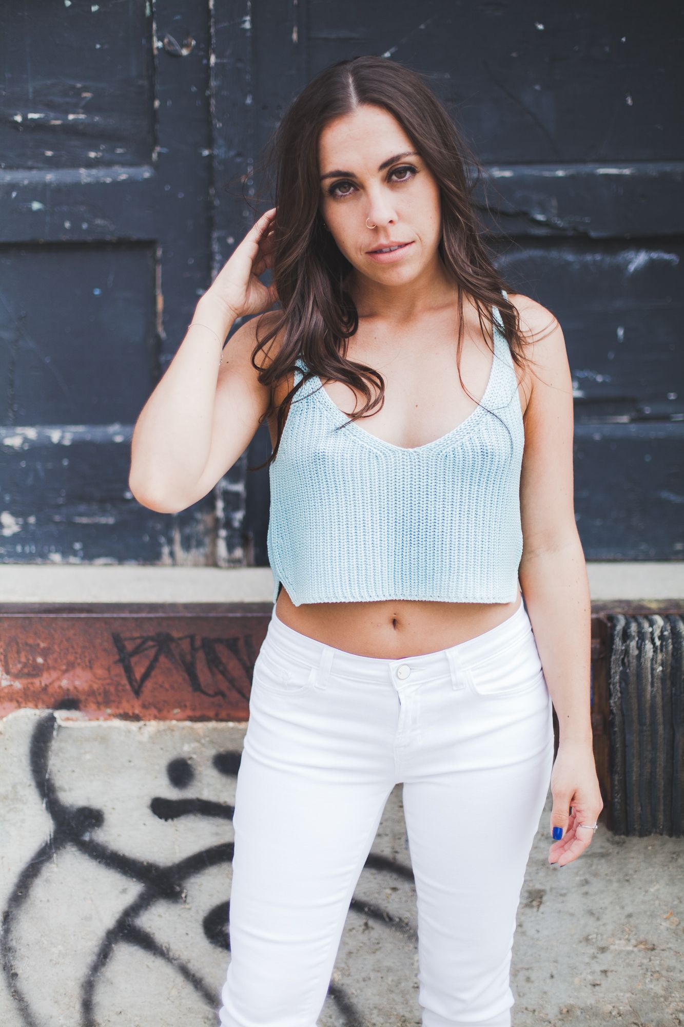 Confident brunette woman in light blue crop top and white jeans posing near a graffiti wall.