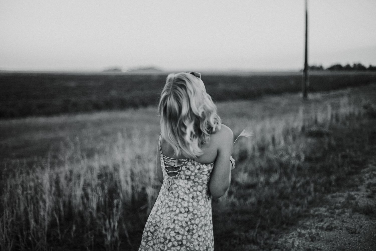 Blonde woman in floral dress standing in a vast green field viewed from behind in black and white.