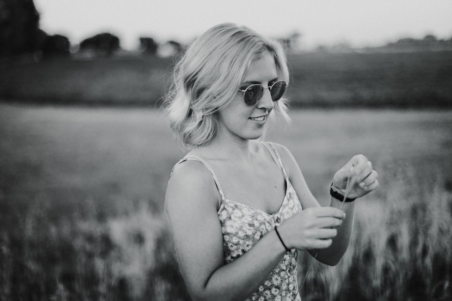 Smiling blonde woman wearing sunglasses and floral dress holding a flower in a scenic field at dusk.