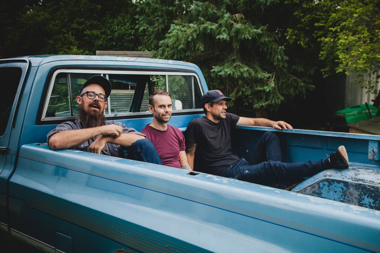 Three men relaxing casually in the bed of a vintage blue pickup truck outdoors.