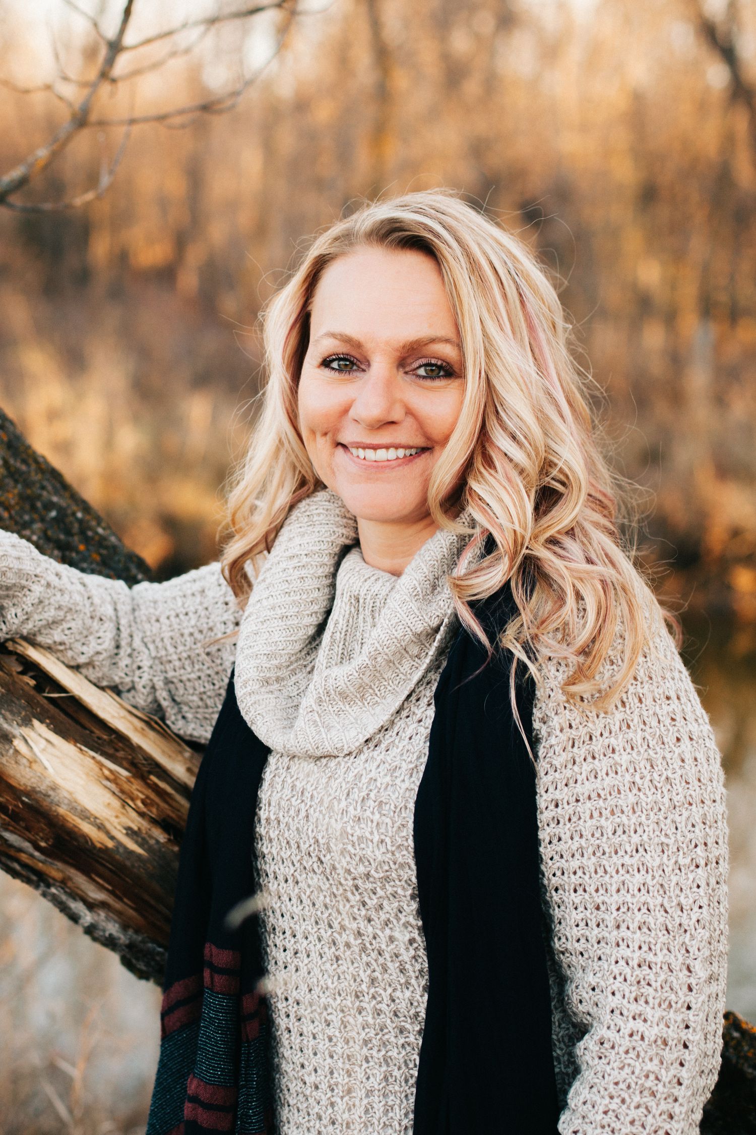 Smiling blonde woman in cozy knit scarf leaning against a tree in autumn outdoor setting.