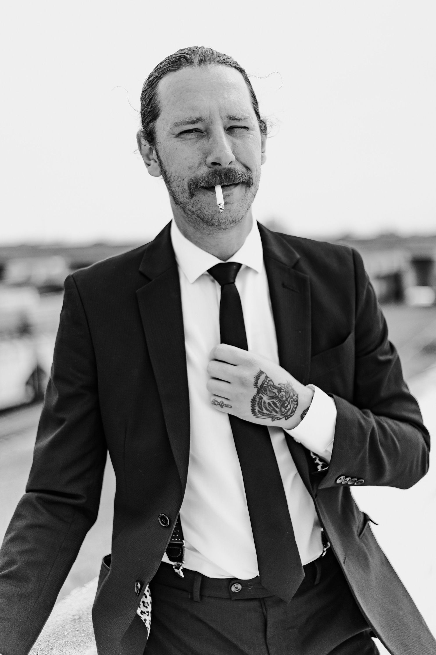 Stylish man in black suit with tattoos and cigarette posing confidently in black and white portrait.