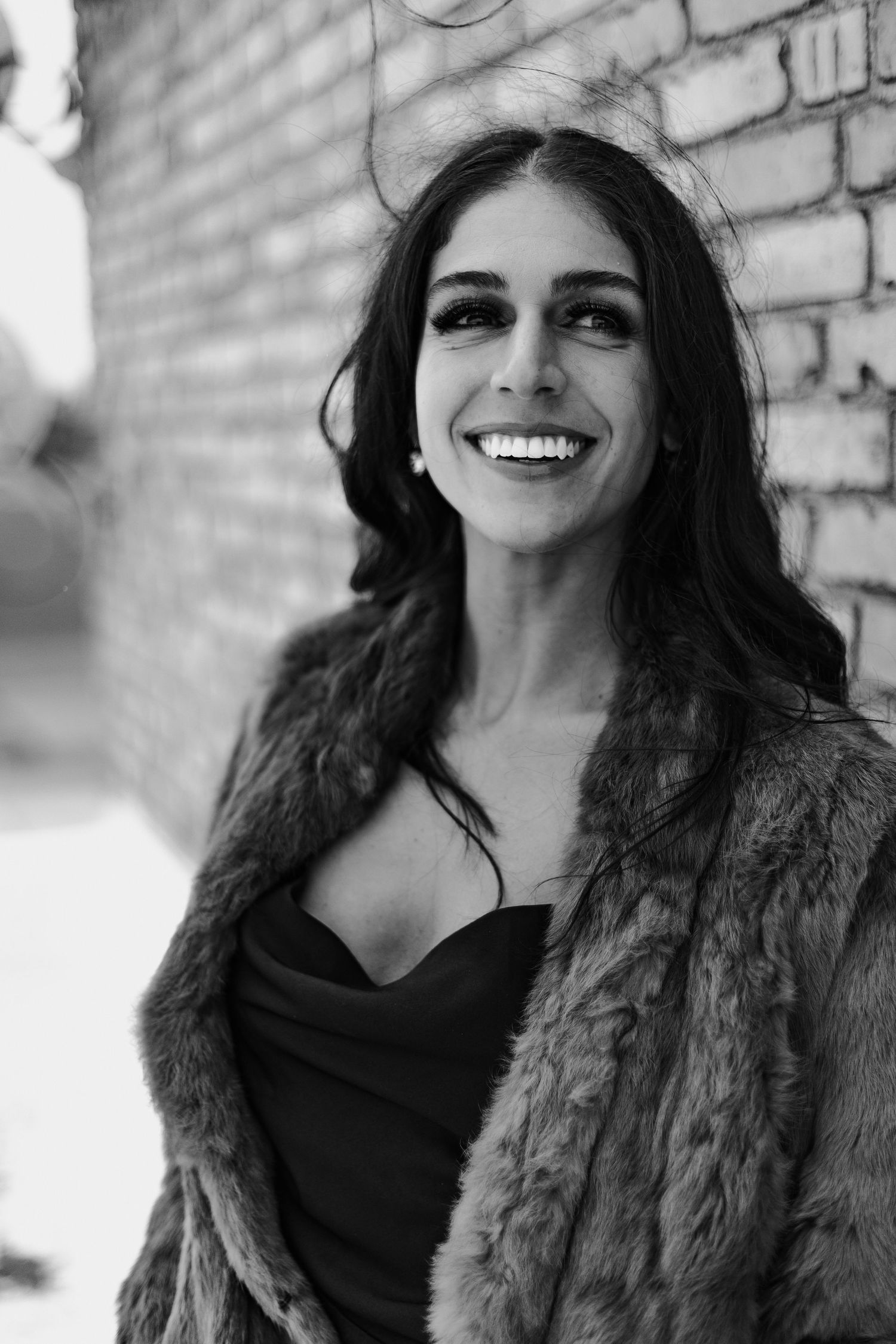Glamorous woman in fur coat laughing against brick wall in stylish black and white portrait photo.