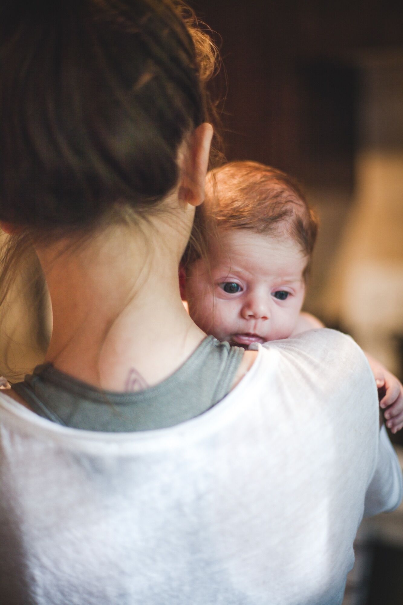 Mother with tattoo gently holding newborn baby against her shoulder in warm indoor lighting.