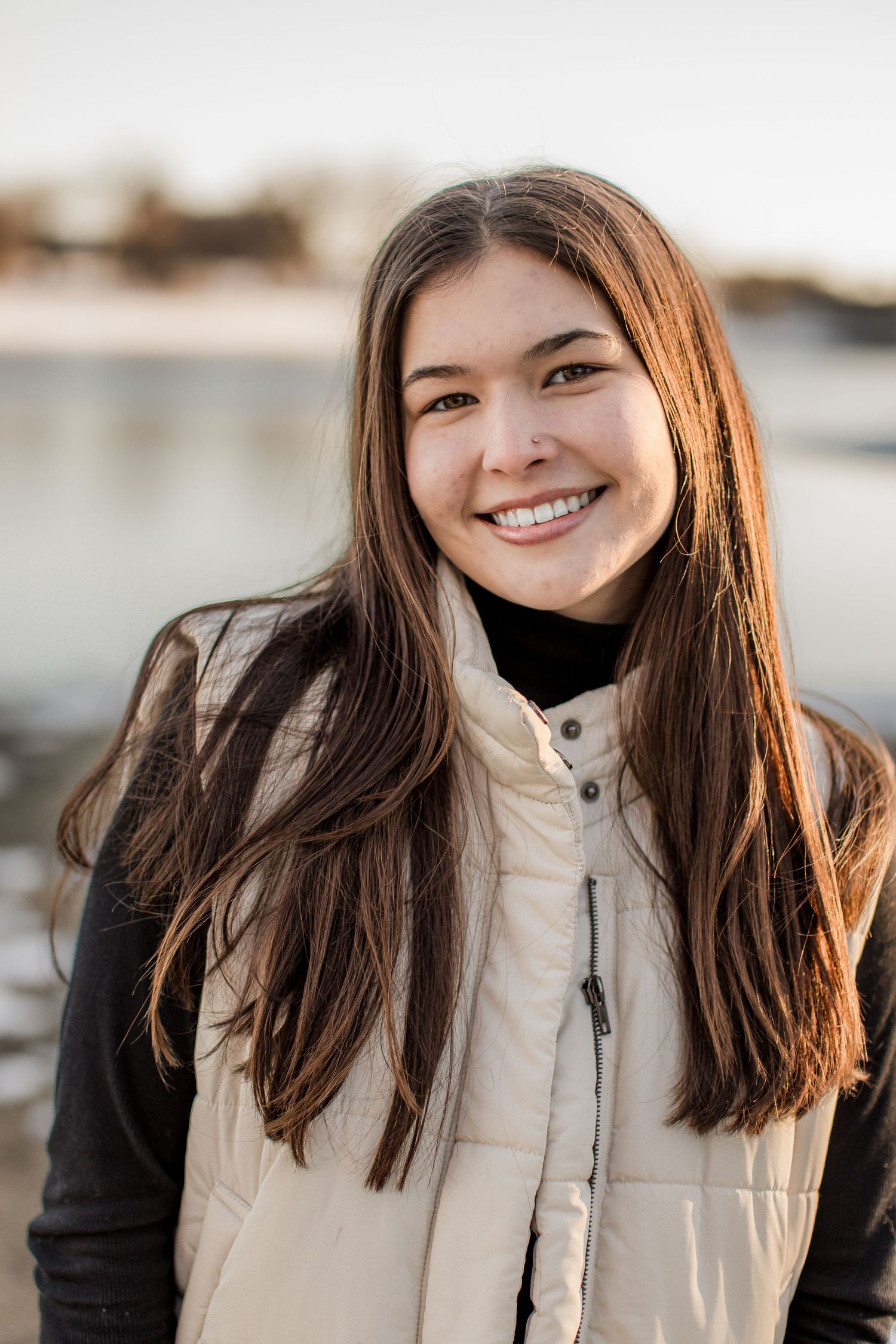 Young woman smiling outdoors near water wearing a cream puffer vest over black turtleneck on a sunny day.