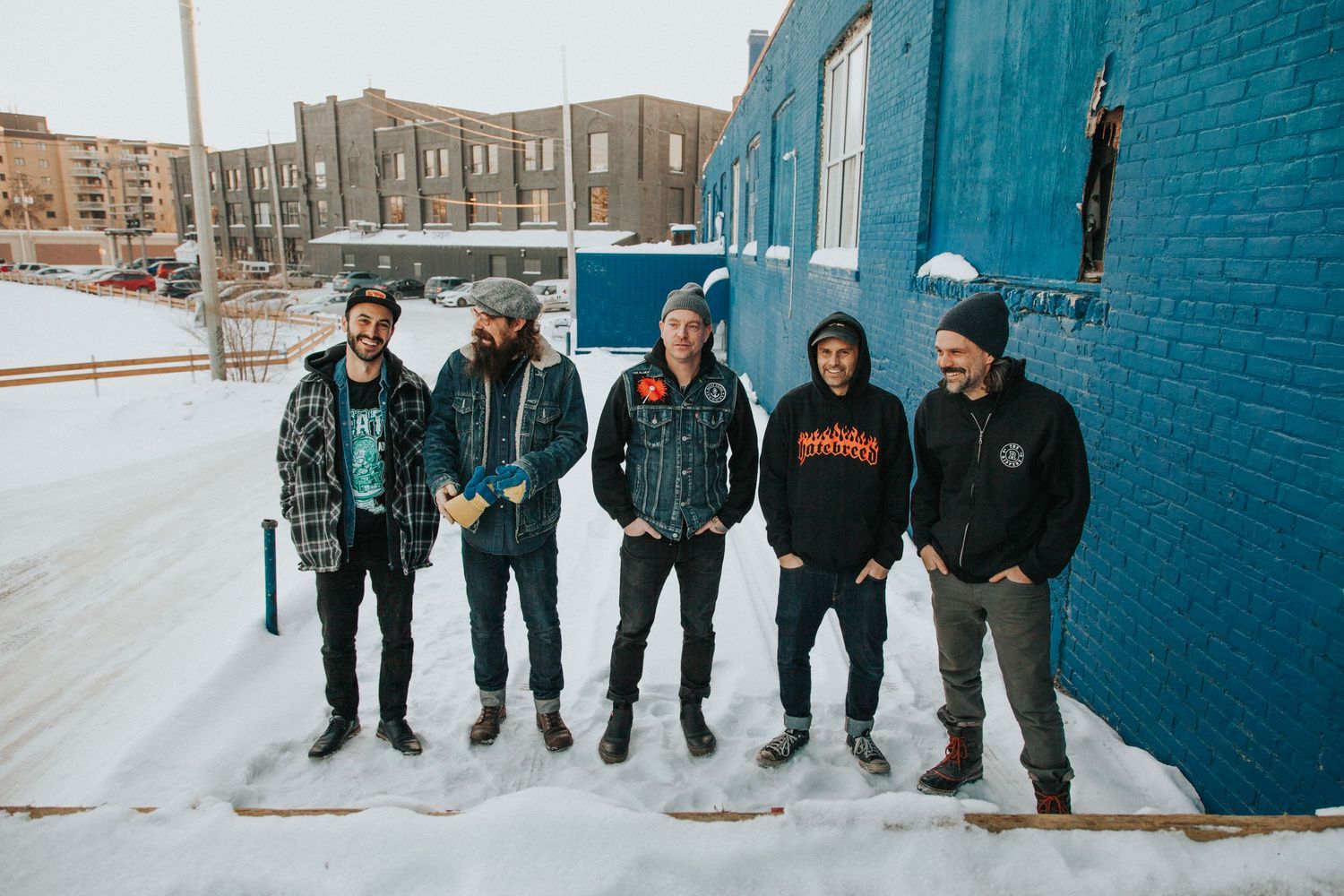 Five friends pose together outdoors in winter snow against a blue brick wall.