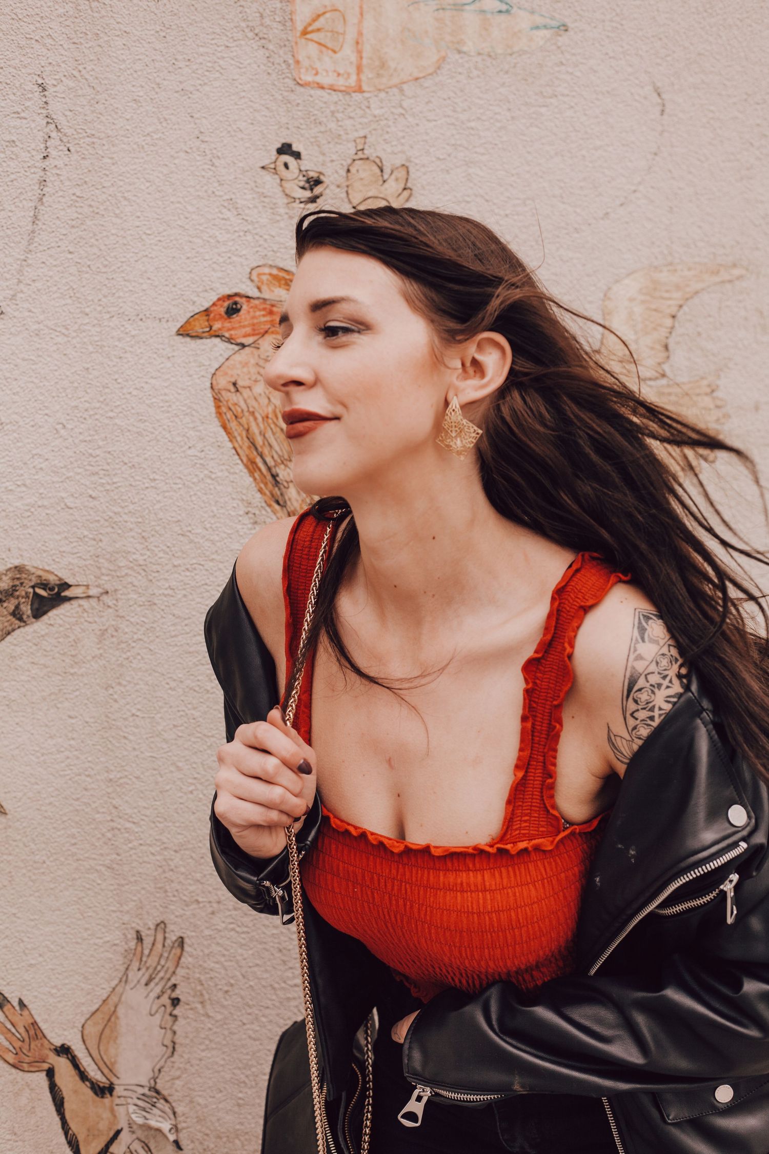 Stylish woman in red top and leather jacket posing against a wall with bird murals.