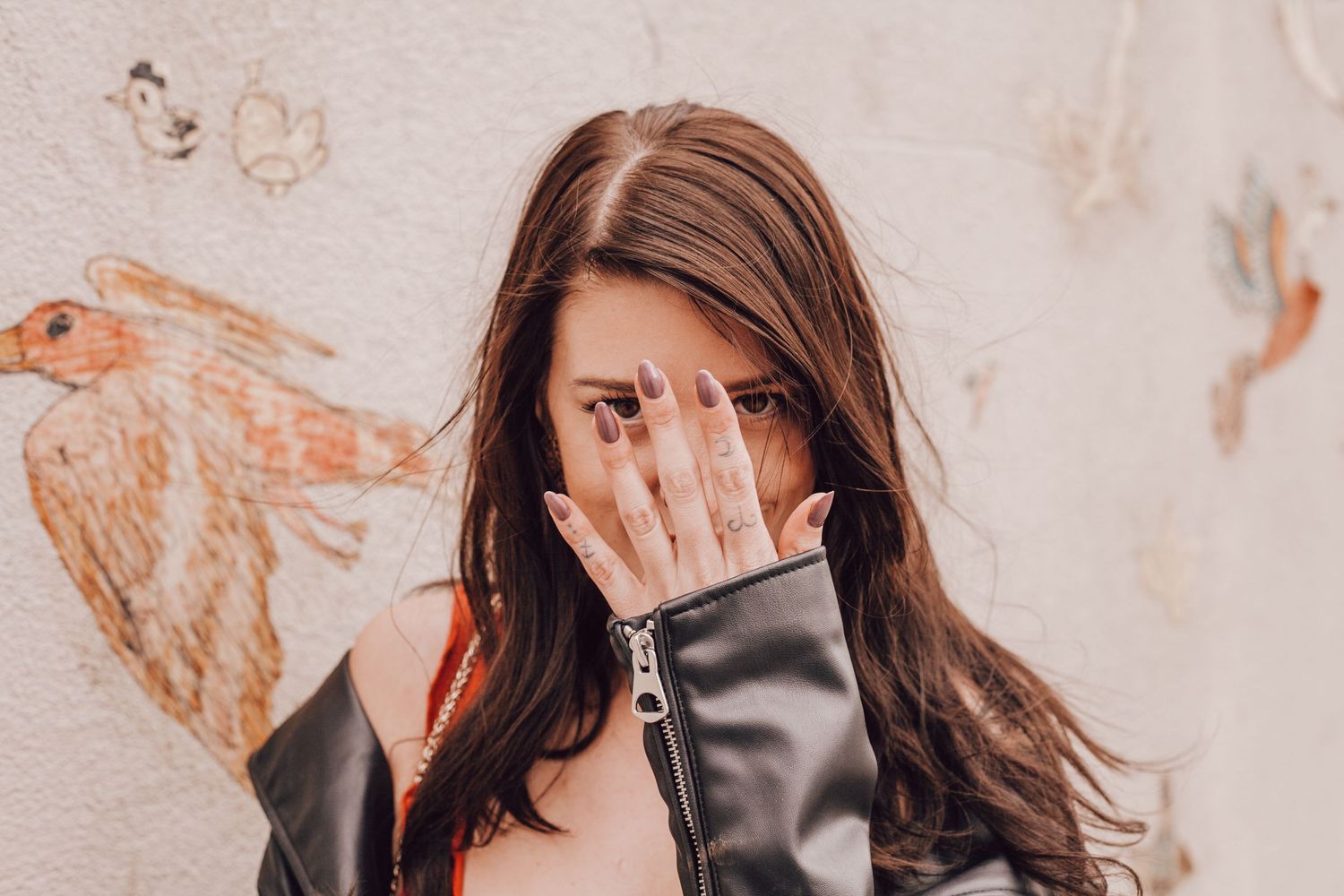 Brunette woman in leather jacket covering her face with her hand against a mural wall.