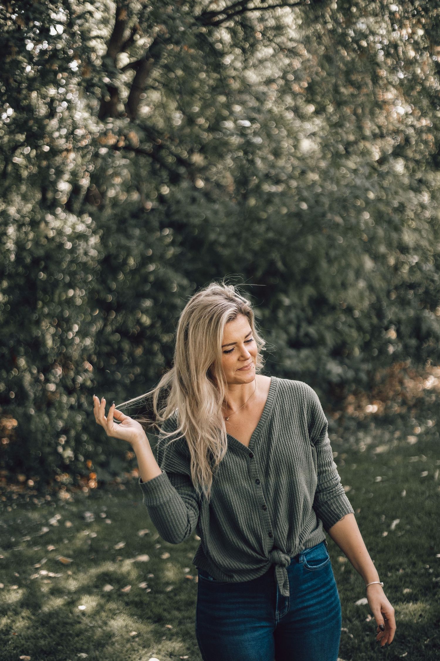 Blonde woman in green top playing with hair in lush garden setting.