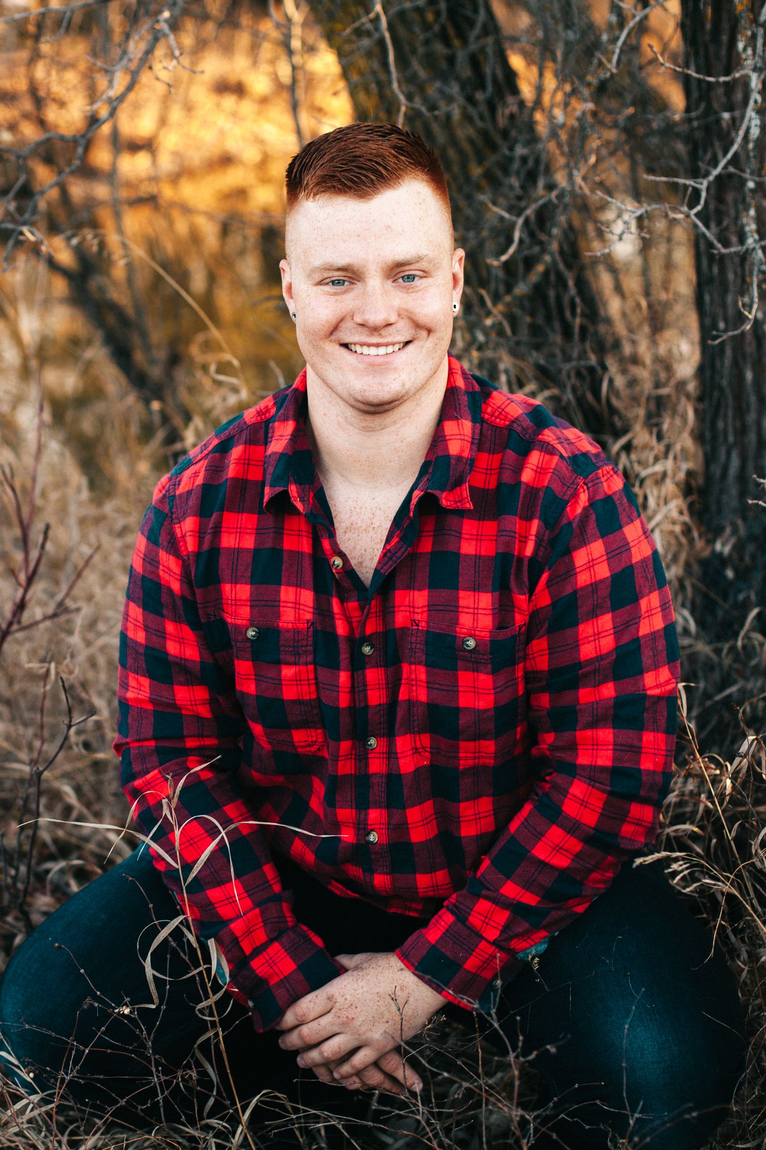 Smiling young man in red plaid flannel shirt posing outdoors among bare branches at sunset.