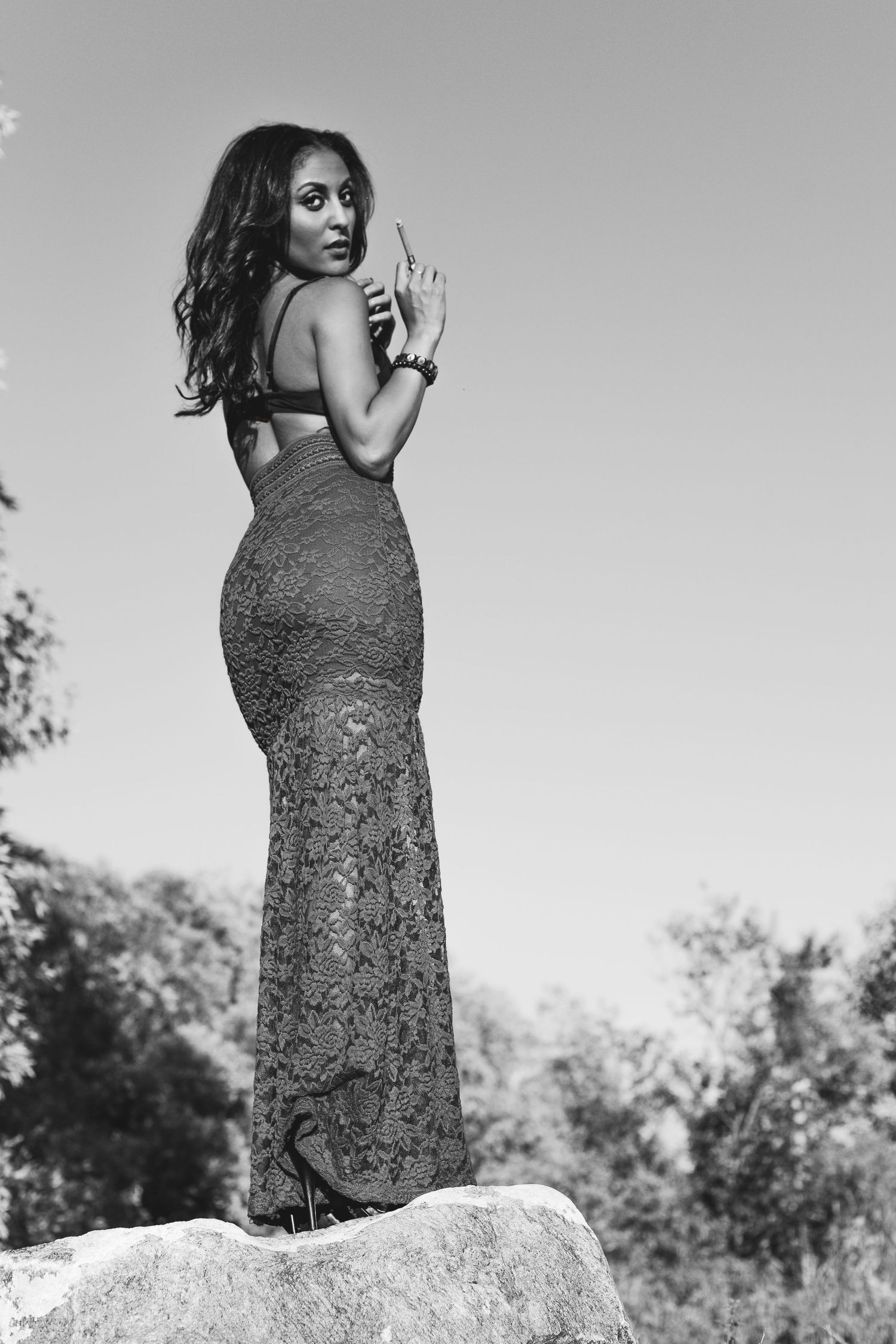 Elegant woman in backless lace gown standing on rock outdoors in dramatic black and white fashion photo.