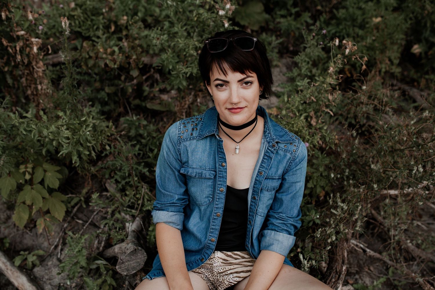 Young woman with short dark hair in denim jacket sitting outdoors among green foliage.