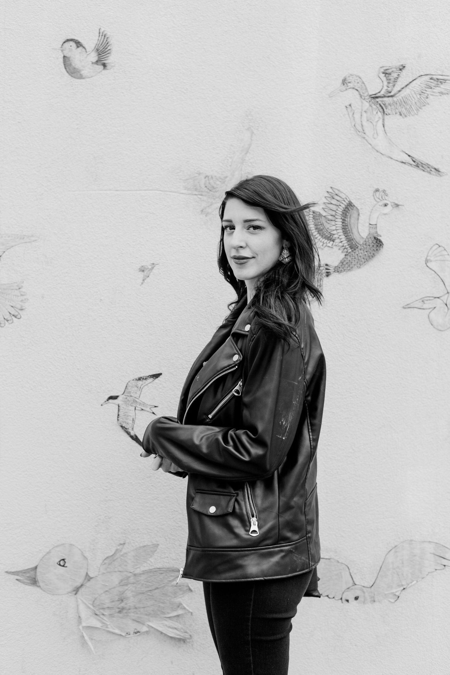 Young woman in a leather jacket posing by a wall with bird artwork in black and white.