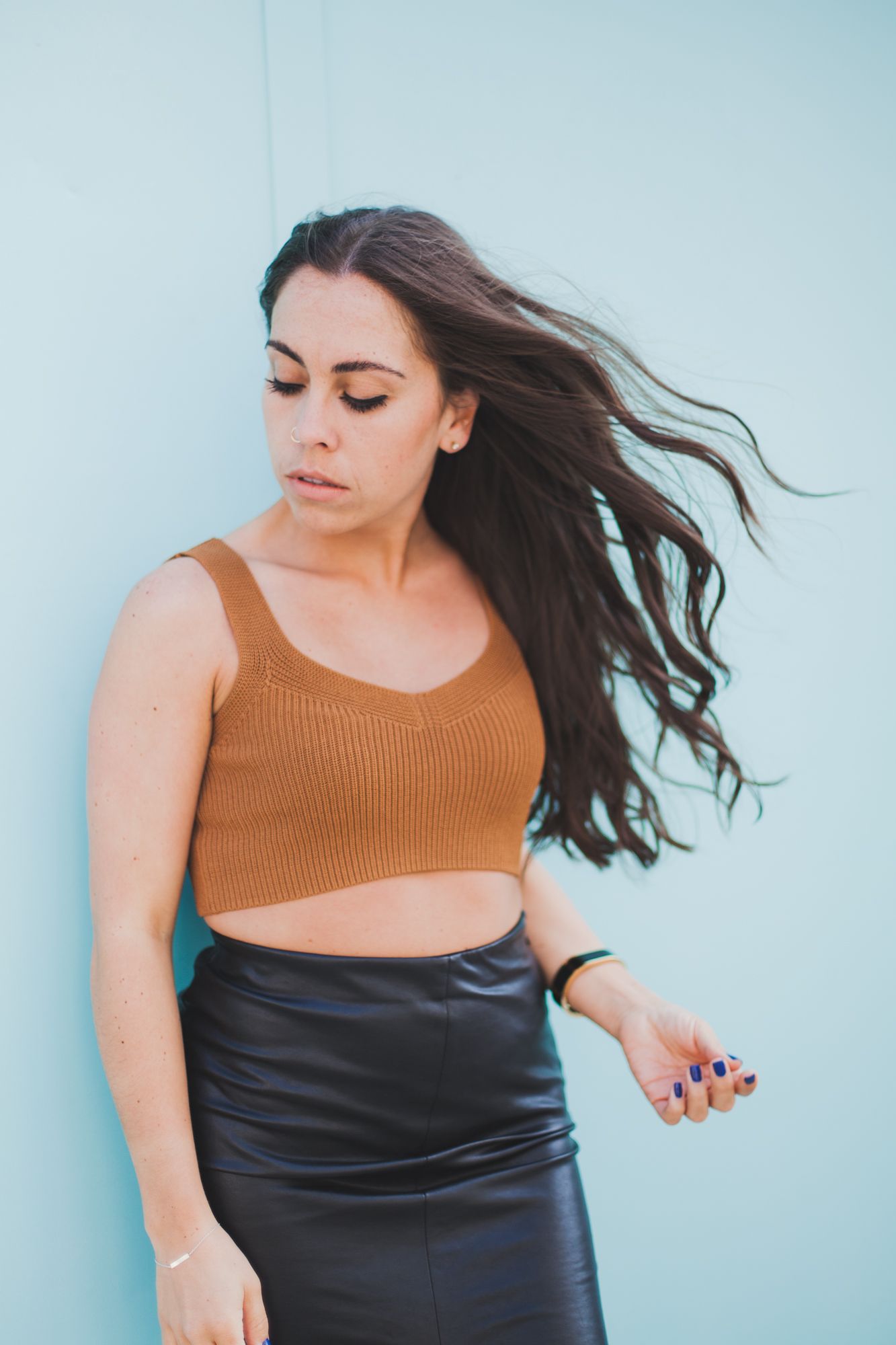 Brunette woman in brown crop top and black leather skirt with wind-blown hair against a teal wall.